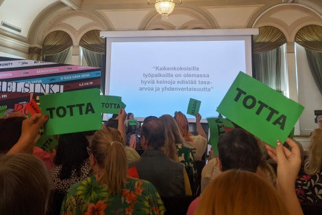 Pride-seminaarin yleisö on nostanut ilmaan vihreitä totta-lappuja, taustalla näkyy teksti: kaikenkokoisilla työpaikoilla on olemassa hyviä keinoja edistää tasa-arvoa ja yhdenvertaisuutta.